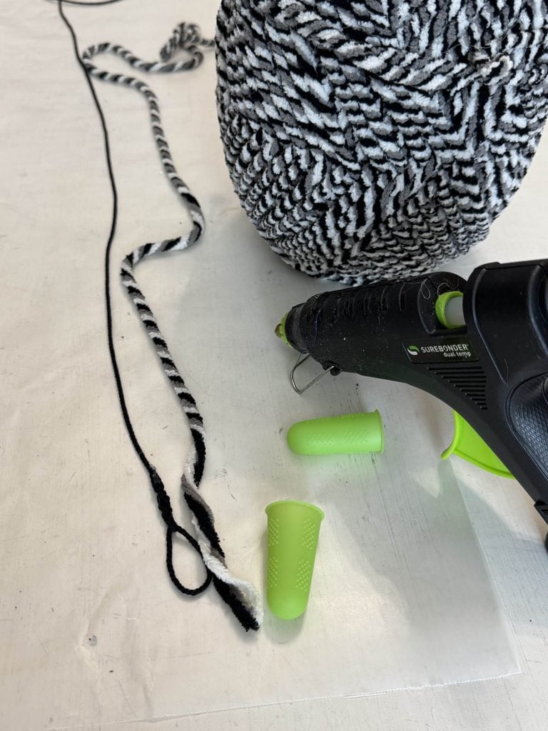 A black and white yarn ball, a twisted cord, a black hot glue gun, and two green silicone finger protectors are on a white surface.
