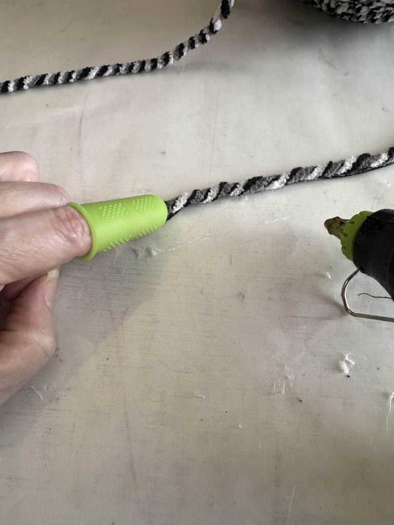 A hand wearing a green finger protector presses a black and white twisted rope onto a surface next to a hot glue gun.
