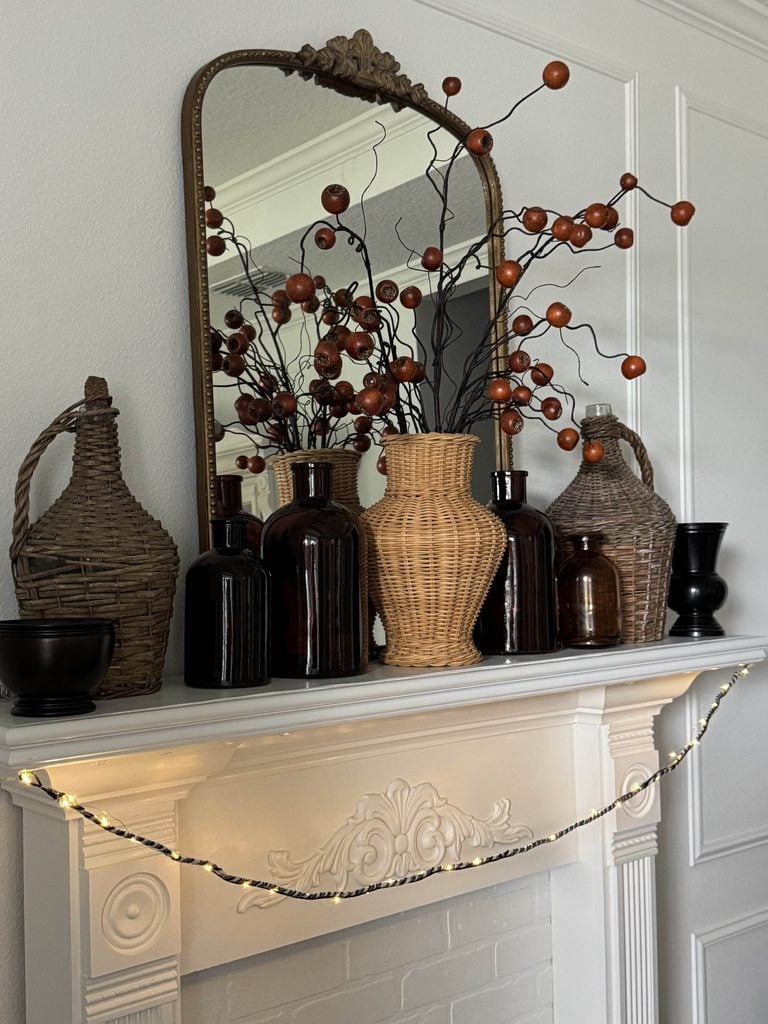 A white mantel decorated with wicker and glass vases, brown bottles, black bowls, and a mirror reflecting a branch arrangement with orange berries. String lights are draped across the mantel.