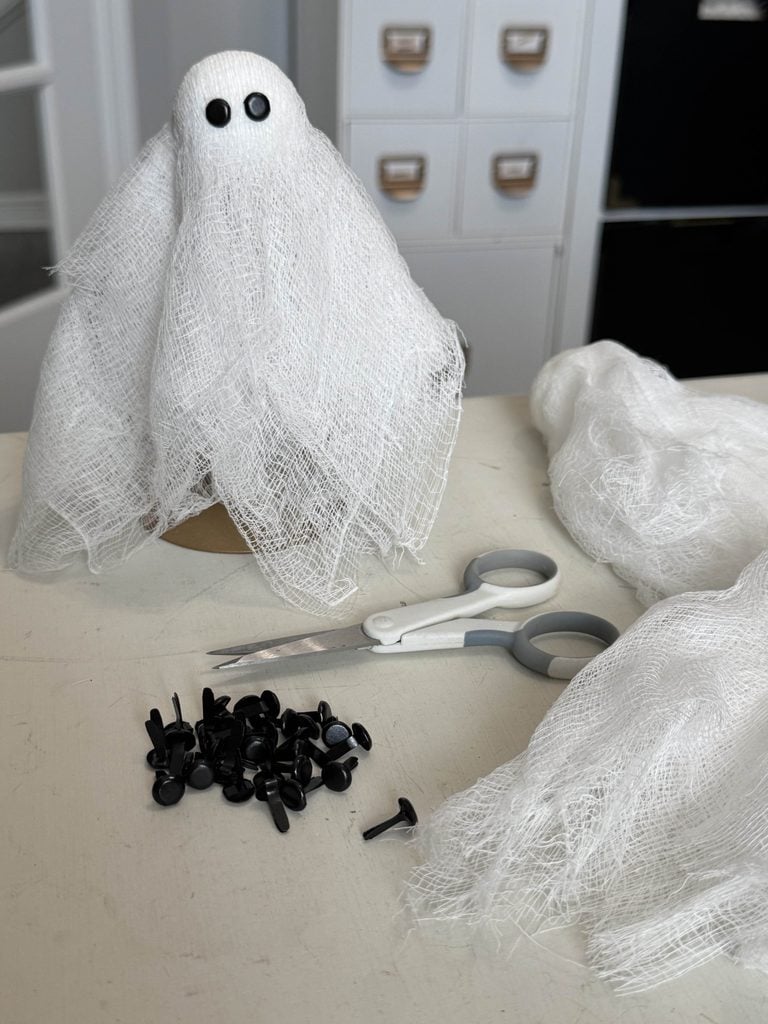 A DIY ghost made from white gauze with black button eyes sits on a table beside scissors, black fasteners, and more gauze. White cabinets and drawers are seen in the background.