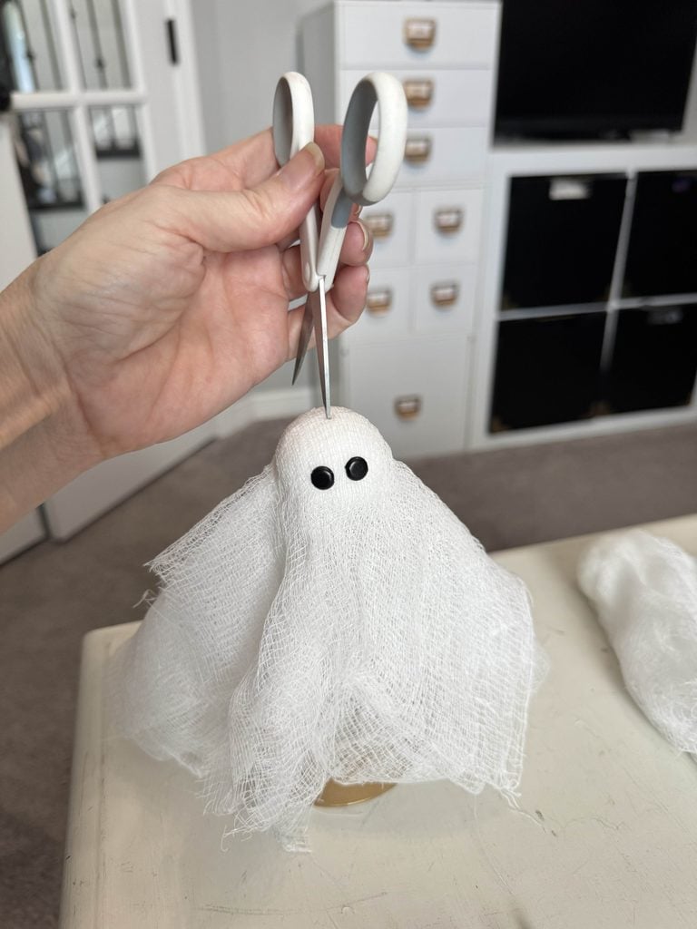 A hand holds scissors above a small ghost decoration made from white gauze with two black eyes, set on a table in a craft room.