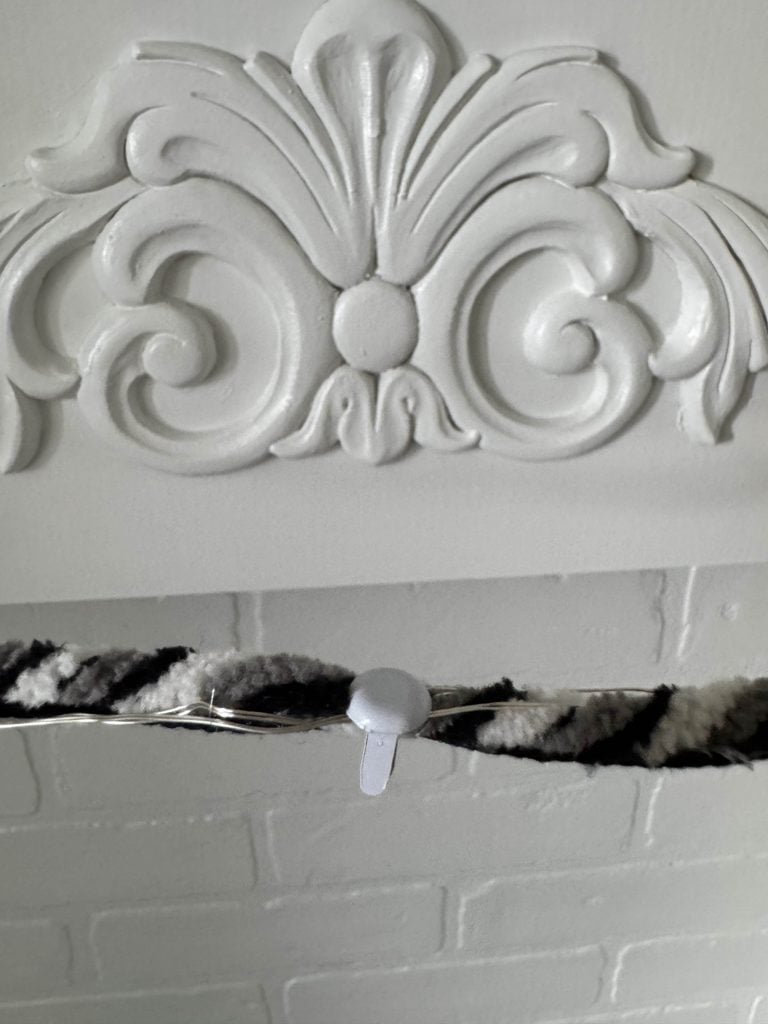 A striped fuzzy garland is attached to a white-painted brick wall using a clear adhesive hook beneath an ornate, white decorative molding.
