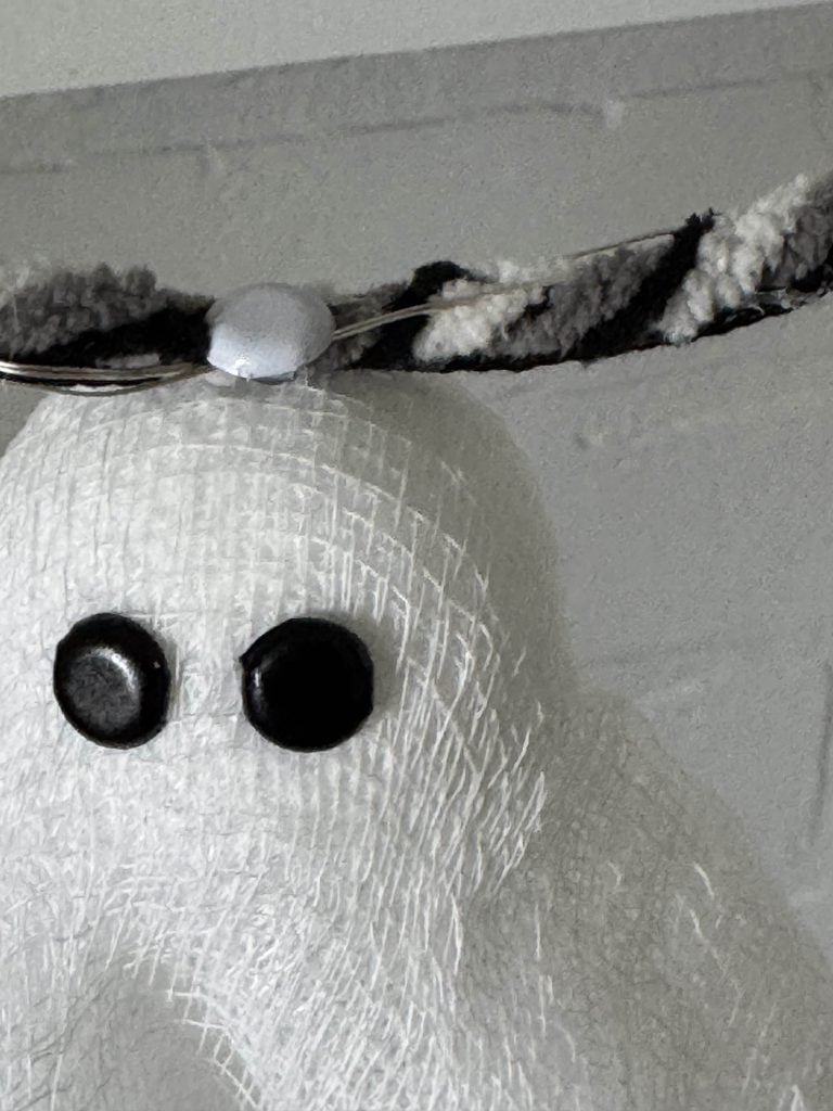 A close-up of a homemade ghost decoration with black button eyes, covered in white gauze, and wearing a striped fuzzy headband.