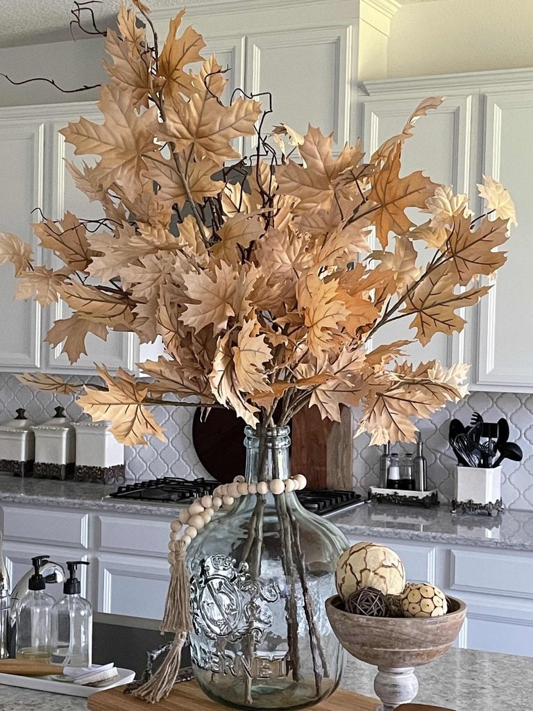 A glass jug filled with dried oak leaves decorates a kitchen island. A wooden bead garland drapes around the jug, and a wooden bowl with decorative balls sits beside it. White cabinets and containers are in the background.
