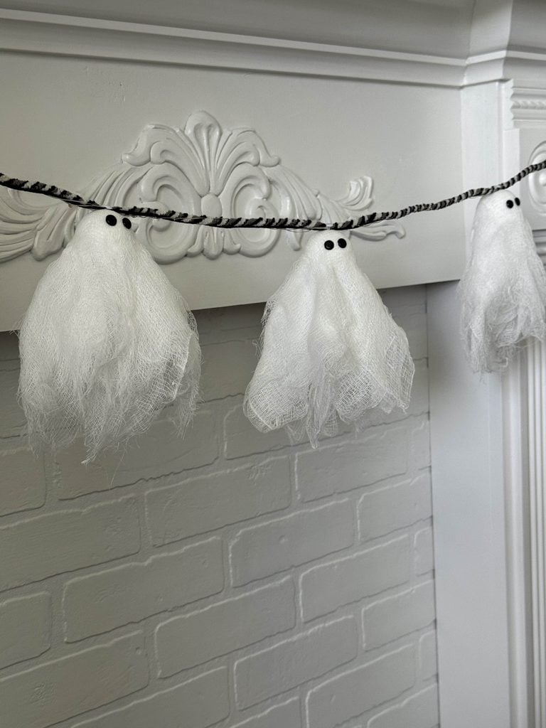 A DIY ghost garland decoration made from cheesecloth, hangs in front of a white brick and ornate mantel.