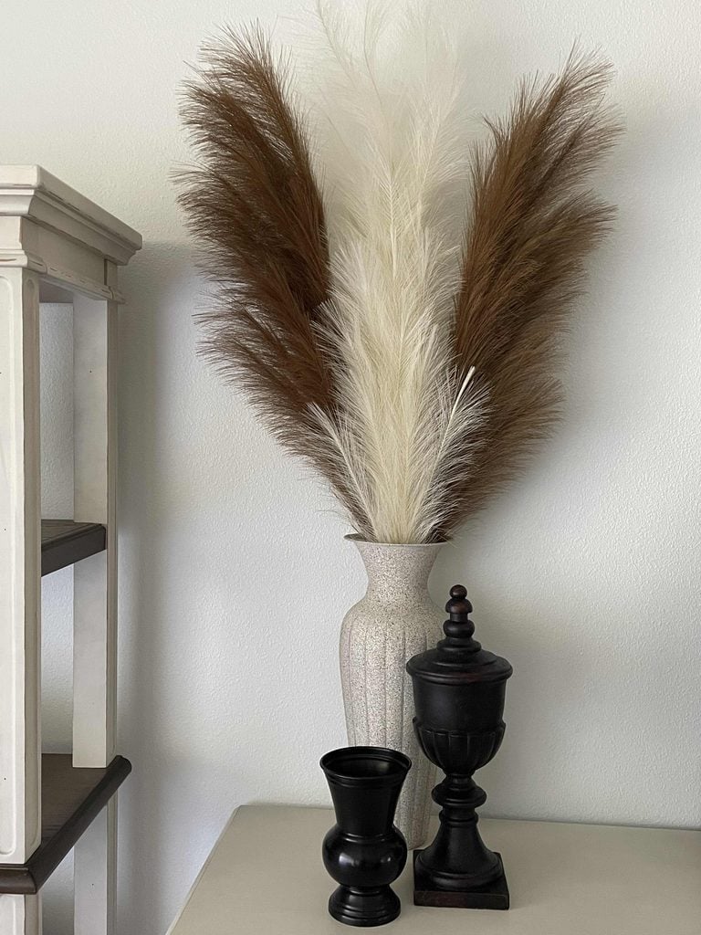 A tall vase with brown and white pampas grass is on a table beside two black decorative objects; neutral-toned furniture is in the background.