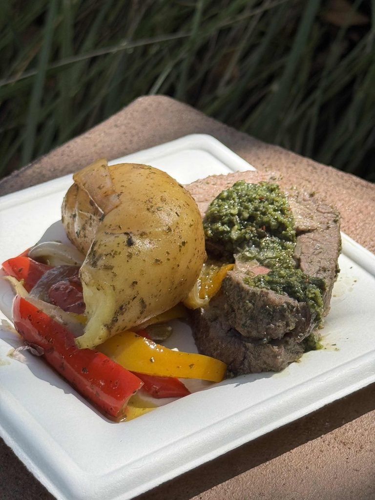 A baked potato, bell pepper strips, and a slice of beef topped with green pesto are served on a white square plate outdoors, with greenery in the background..