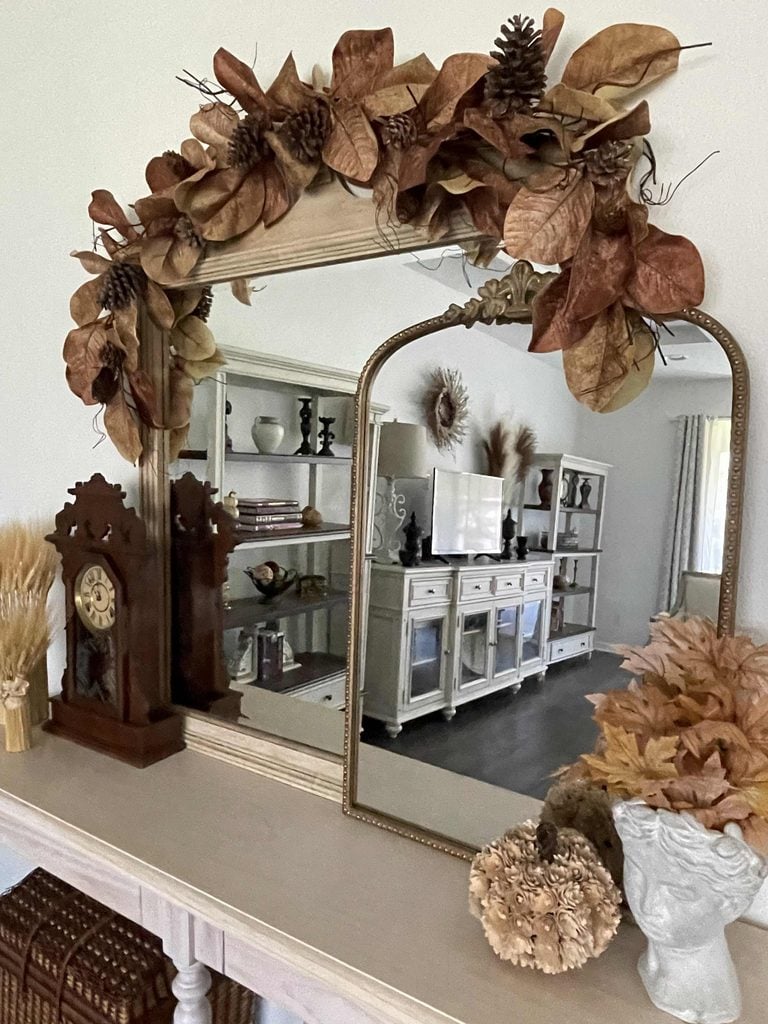 A mirror decorated with dried leaves and pinecones reflects a living room. On the mantle are a clock, wheat stalks, and autumn-themed decorations, including a pumpkin and a white vase with dried leaves.
