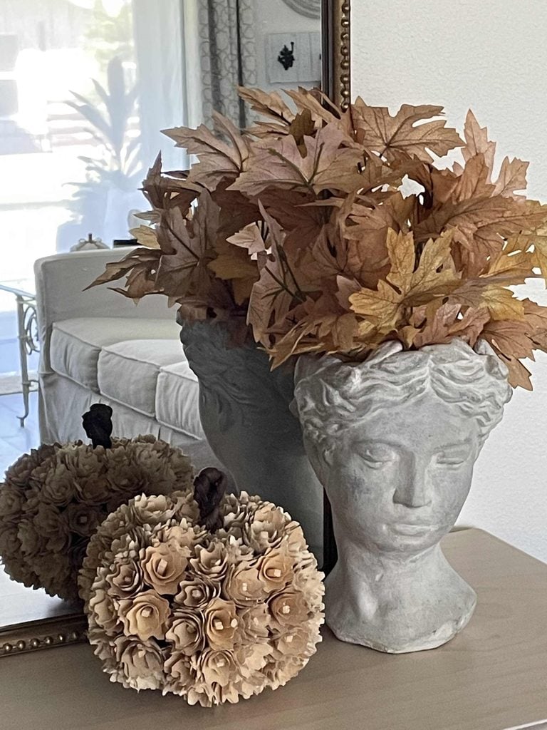 Stone bust vase with dried leaves and two decorative pumpkins made of wooden flowers on a light wood table, reflected in a mirror. An off-white sofa and window can be seen in the background.