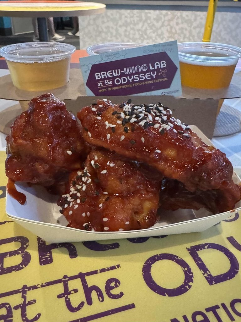 Three saucy chicken wings topped with sesame seeds in a paper tray, with two cups of beer and a Brew-Wing Lab at the Odyssey sign in the background.
