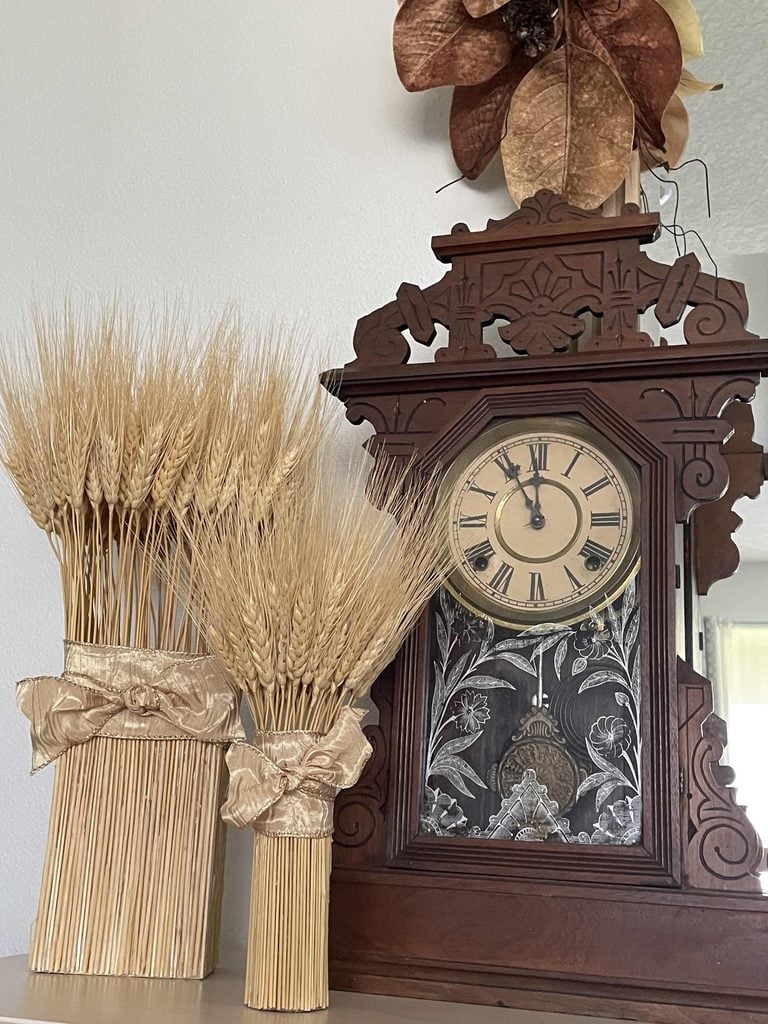 An ornate antique clock with a floral design sits next to two decorative bundles of dried wheat tied with ribbon; a dried flower arrangement is on top of the clock.