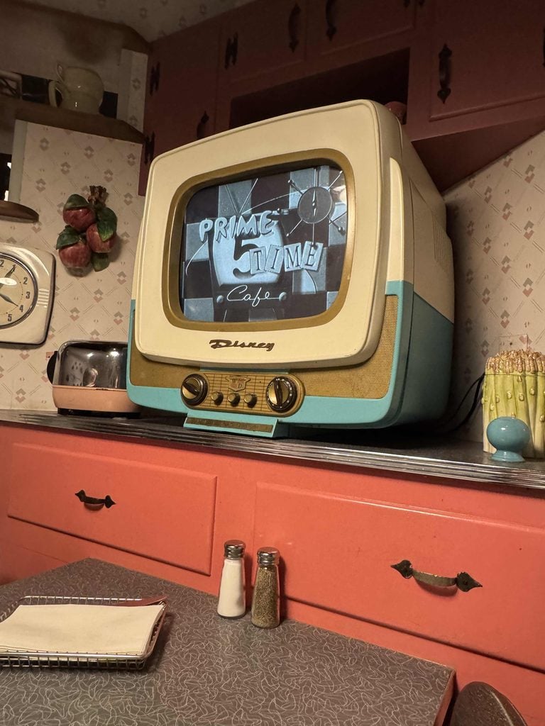 A retro Disney TV on a vintage kitchen counter displays Prime Time Cafe in a scene that radiates Seasonal Sentiments. Nearby are a notebook, salt and pepper shakers, a napkin holder, a clock, and nostalgic kitchen decor.