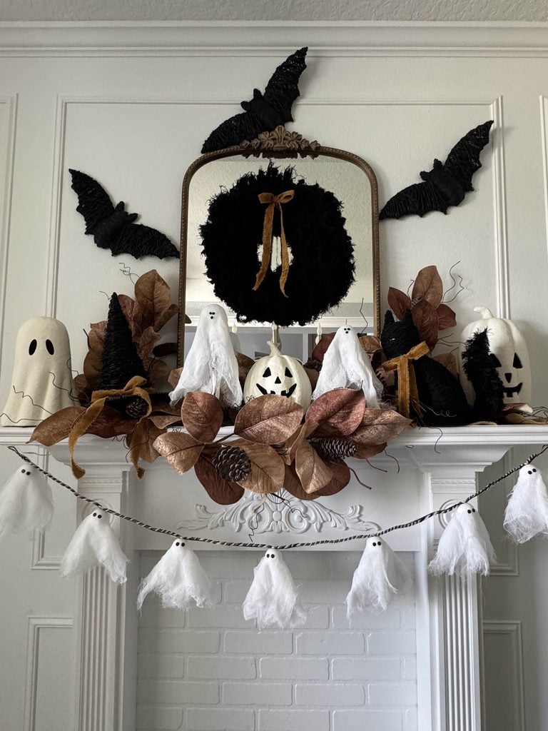 A Halloween mantel decor scene features faux leaves, pumpkins, ghost and bat decorations, a mirror, and a garland of small ghosts hanging below the shelf.