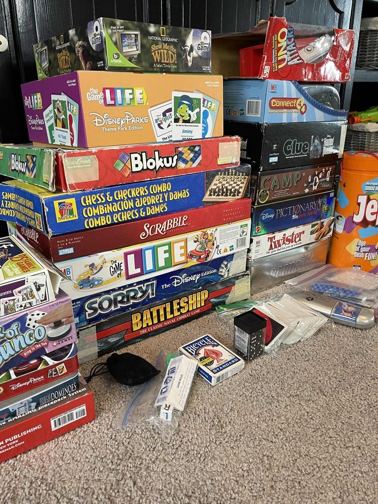 A stack of assorted board games, including Life, Clue, Connect 4, Sorry!, Battleship, and Scrabble, sit on a carpeted floor next to playing cards, dominoes, Jenga, and other game pieces.