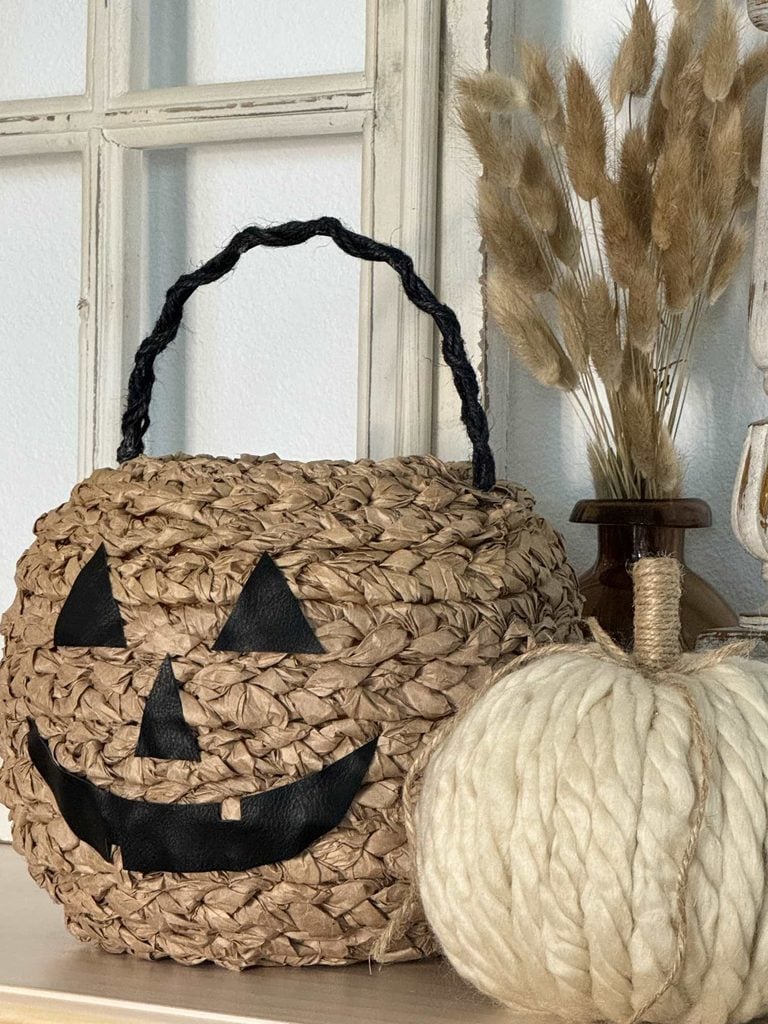 A woven basket shaped like a jack-o-lantern sits next to a white yarn pumpkin and dried grass in a vase, all arranged on a shelf in front of a window—perfect inspiration for anyone looking for creative DIY pumpkin ideas.