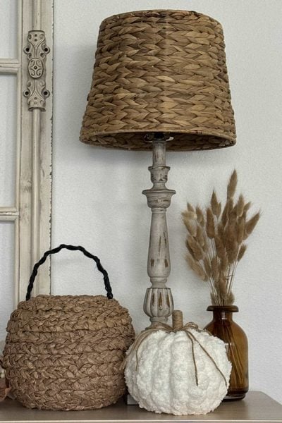 A woven table lamp, a wicker basket, a white textured pumpkin inspired by DIY pumpkin ideas, and a brown vase with dried grass are arranged on a neutral-colored surface by a cream-colored window frame.