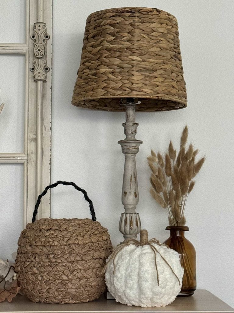 A woven table lamp, a wicker basket, a white textured pumpkin, and a brown vase with dried grass are arranged on a neutral-colored surface by a cream-colored window frame.