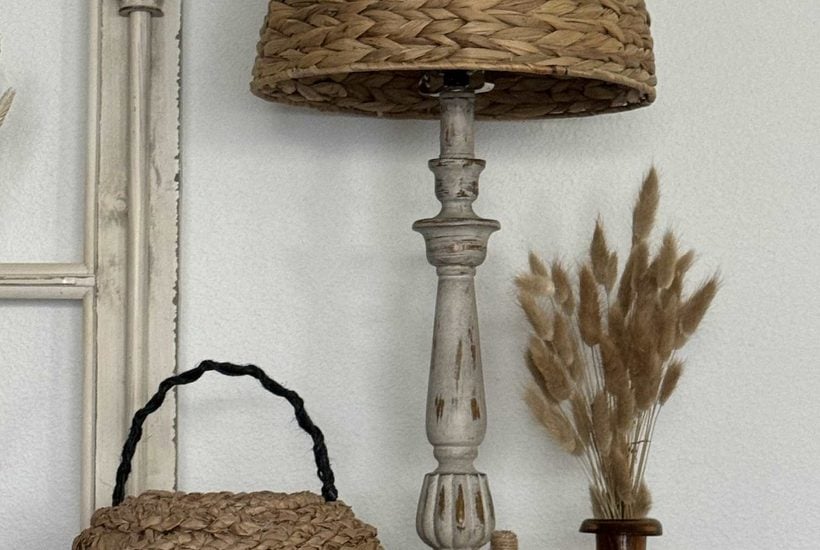 A woven table lamp, a wicker basket, a white textured pumpkin inspired by DIY pumpkin ideas, and a brown vase with dried grass are arranged on a neutral-colored surface by a cream-colored window frame.