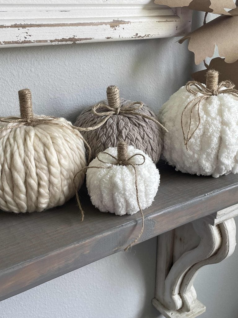 Four decorative pumpkins made from soft, chunky yarn in cream, gray, and white tones sit on a wooden shelf. Each pumpkin has a twine-wrapped stem and a simple twine bow, creating a cozy fall display.