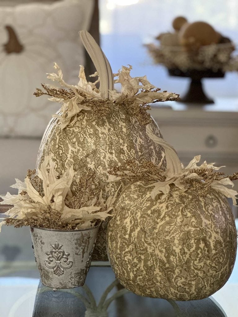Three decorative pumpkins with intricate beige patterns and cream-colored leaves—perfect for DIY pumpkin ideas—are arranged on a glass table. One is placed in a small ornate pot. The soft background includes a white pillow and a tray with additional decor.