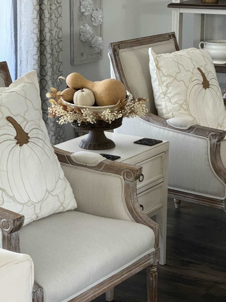 Two cream-colored armchairs with DIY pumpkin pillows sit beside a small table. On the table is a pedestal bowl filled with decorative gourds and small pumpkins.