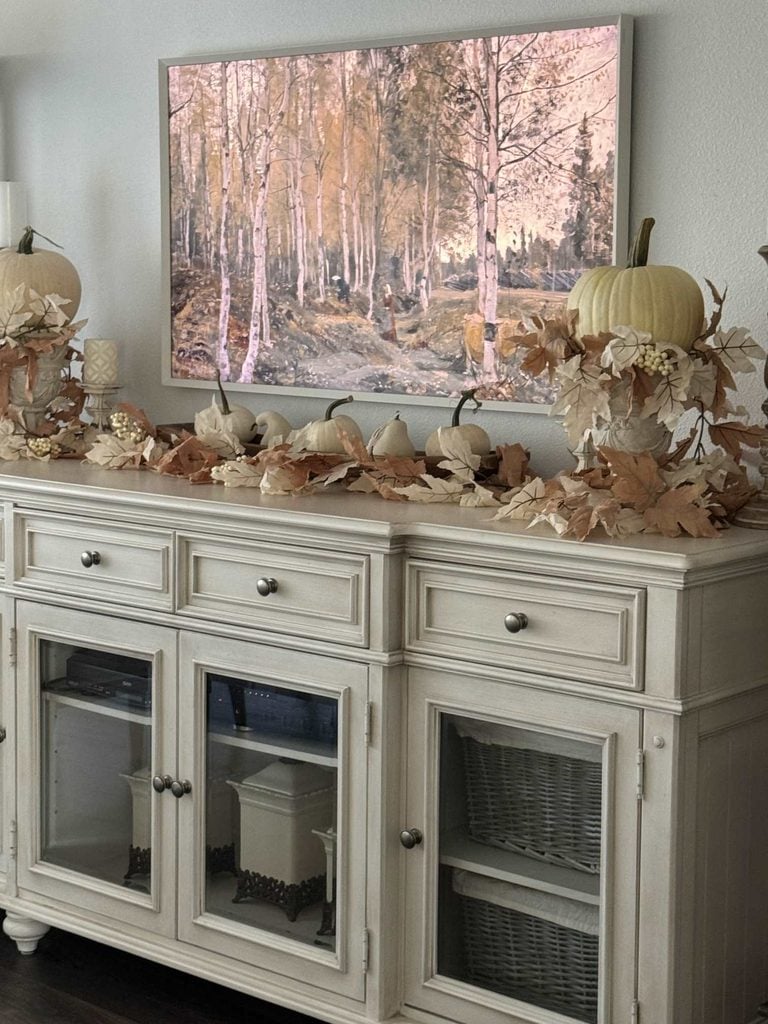 A white sideboard decorated with cream pumpkins, autumn leaves, and candles sits below a wall-mounted painting of a forest with birch trees in fall colors.