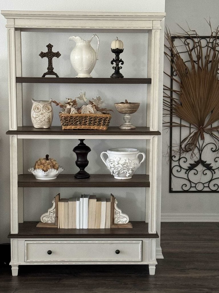 A white bookshelf with various decor items, including a cross, owl, vases, books, a candle, and a basket—thoughtfully arranged to inspire natural fall decor ideas—set against a wall adorned with a decorative metal and dried leaf art piece.