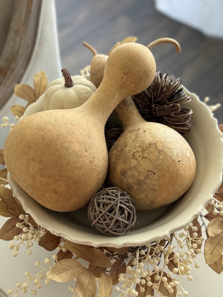 A decorative bowl filled with dried gourds, a white pumpkin, woven balls, and dried foliage, creating an autumn-themed centerpiece.
