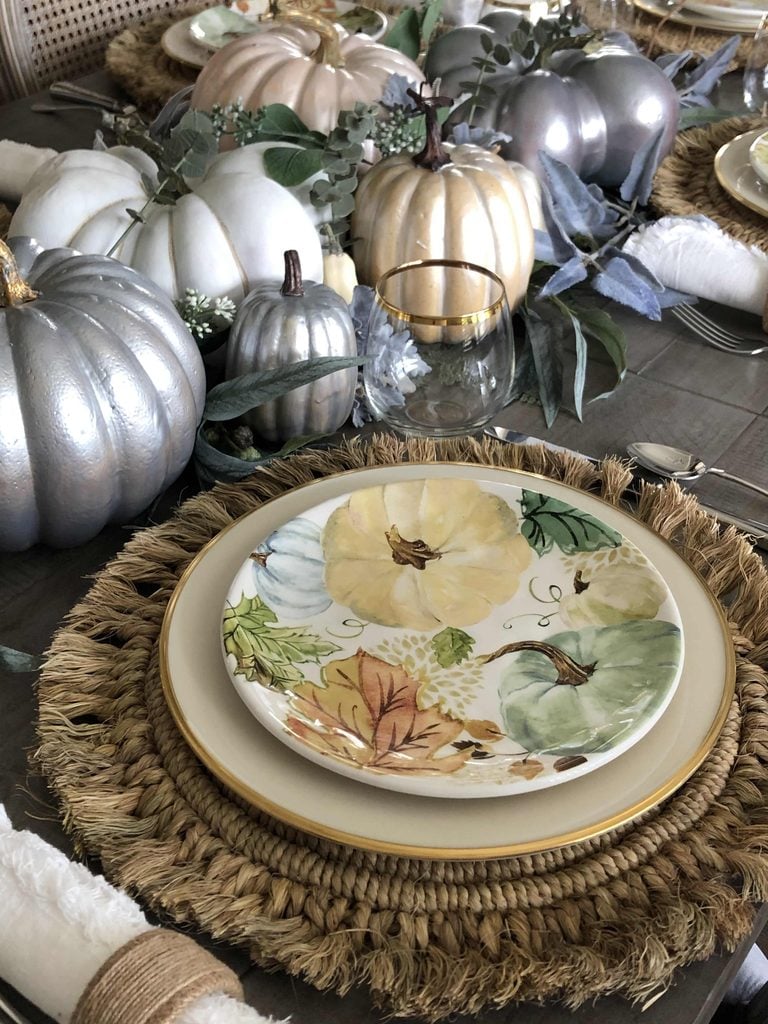A fall-themed table setting with a woven placemat, a decorative pumpkin plate, gold-rimmed glass, and silver, white, and gold pumpkins with greenery as a centerpiece—perfect inspiration for your own DIY pumpkin ideas.