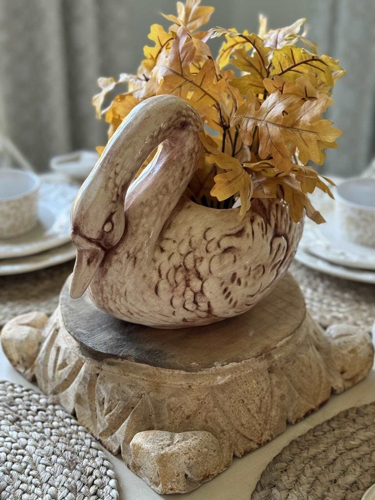 A ceramic swan vase with yellow leaves sits on a carved stone pedestal, surrounded by woven placemats and cream-colored dishes set on a dining table.