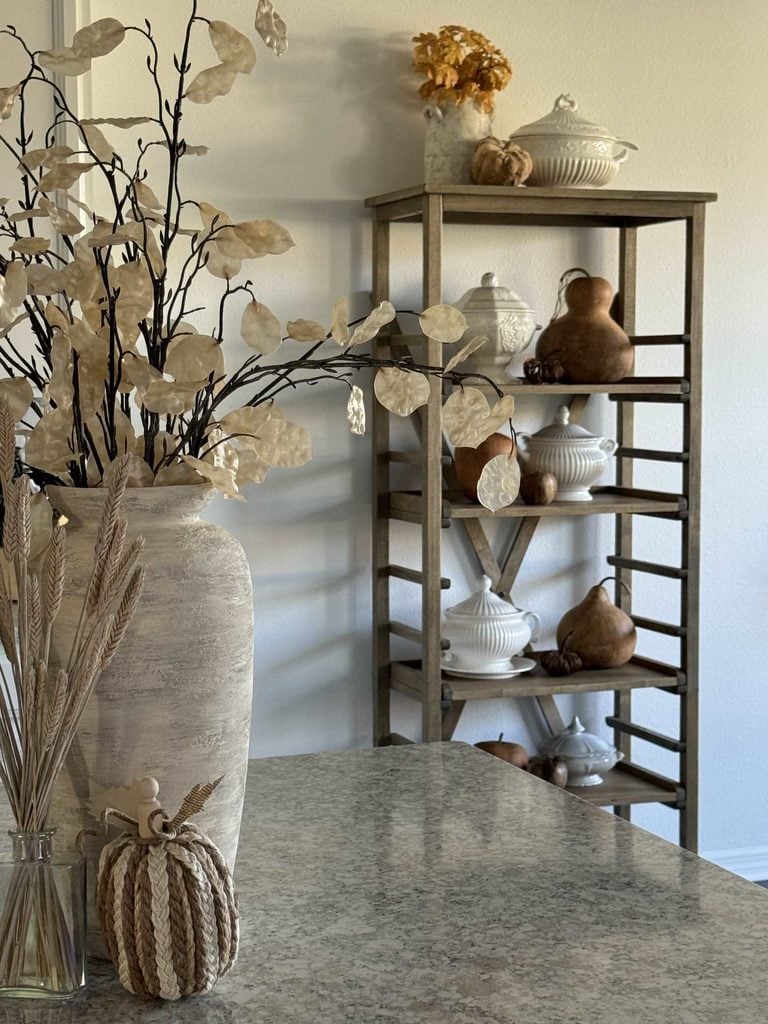 Neutral-toned kitchen with a marble counter, a large white vase holding dried branches, a glass jar with wheat, and a shelf in the background displaying white ceramic dishes and brown decorative gourds.