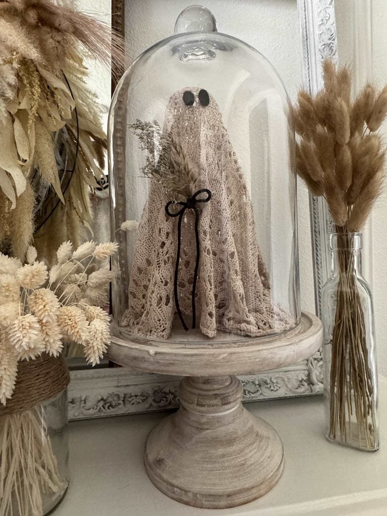 A small ghost figurine with black eyes, draped in lace, stands under a glass dome. It holds dried flowers and is surrounded by more dried floral arrangements on a rustic wooden pedestal.