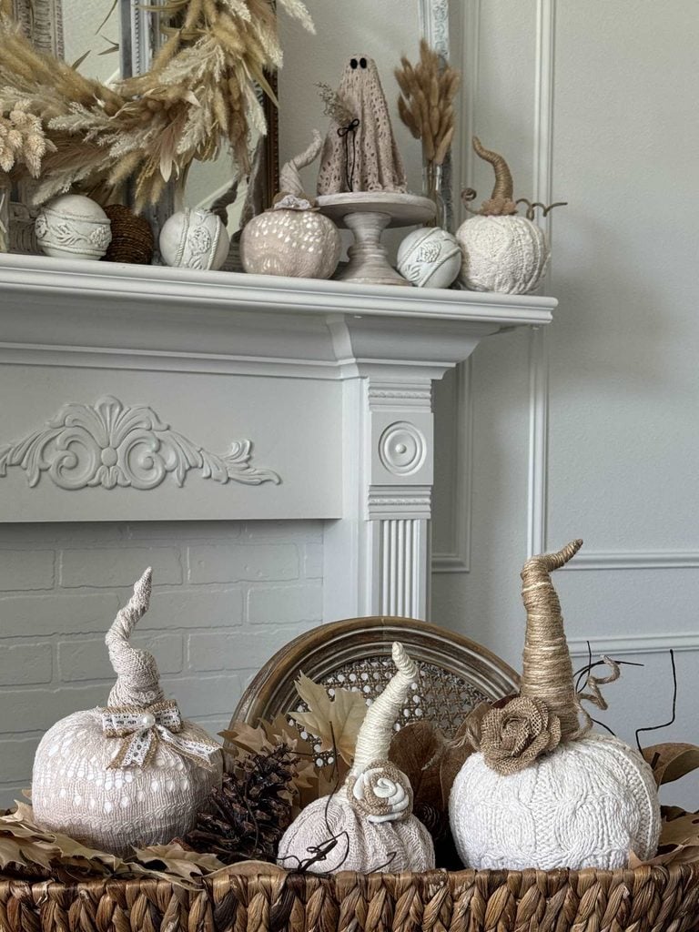 Neutral-toned, textured pumpkins and pinecones in a basket sit before a white fireplace; the mantel displays more pumpkins, dried flowers, and a ghost figurine in a cozy, autumn-themed scene—perfect inspiration for your next DIY pumpkin ideas.