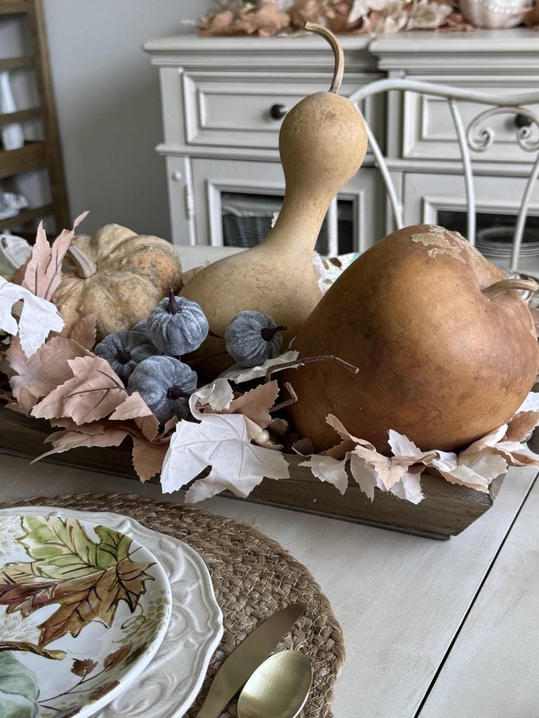 A wooden tray holds gourds, gray berries, and pale leaves on a dining table set with fall-themed plates, woven placemats, and gold utensils. A white hutch and autumn decor are in the background.