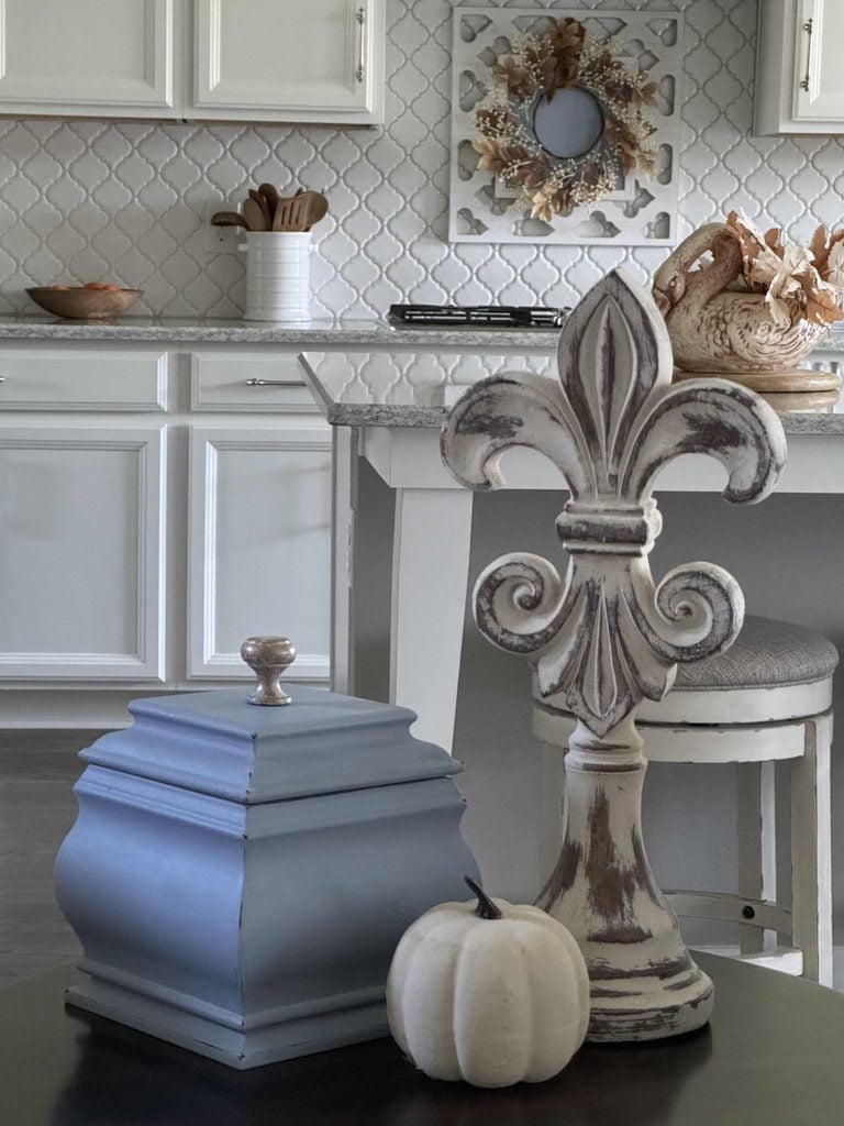 A rustic kitchen features a fleur-de-lis statue, a blue decorative box, and a small white pumpkin on a table—perfect natural fall decor ideas—while white cabinets and neutral decor provide a charming backdrop.