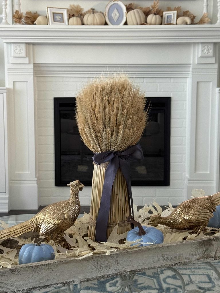 A bundle of wheat tied with a dark ribbon, flanked by gold bird figurines and blue pumpkins sits on a table in front of a white fireplace decorated with more pumpkins and framed photos.