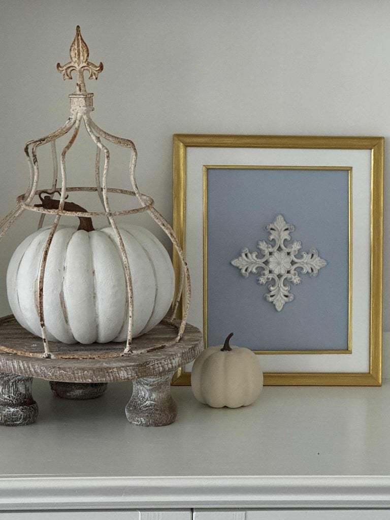 A white pumpkin sits on a rustic wooden stand with a decorative metal cage. Next to it is a small cream pumpkin and a framed ornate design with a gold border on a gray background.
