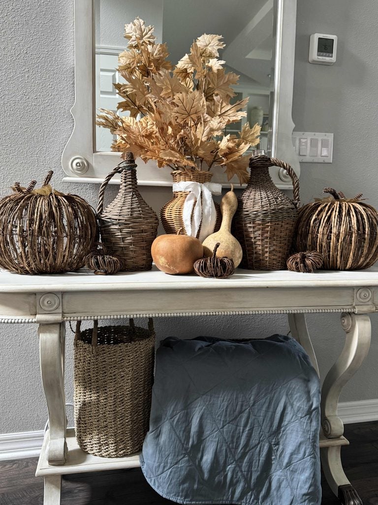 A beige console table with woven baskets, dried leaves in a vase, gourds, and wicker pumpkins sits against a gray wall with a mirror above. A basket and folded blue blanket are placed underneath the table.