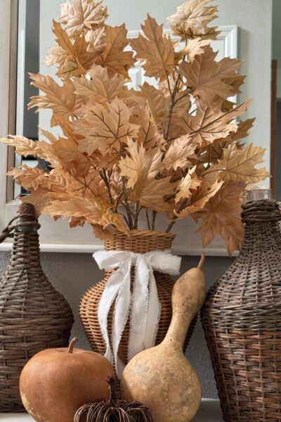 A rustic fall arrangement featuring dried golden leaves in a wicker vase with a white ribbon, flanked by woven bottles, a gourd, a pumpkin, and a small brown decorative item on a mantel—perfect inspiration for those seeking natural fall decor ideas.