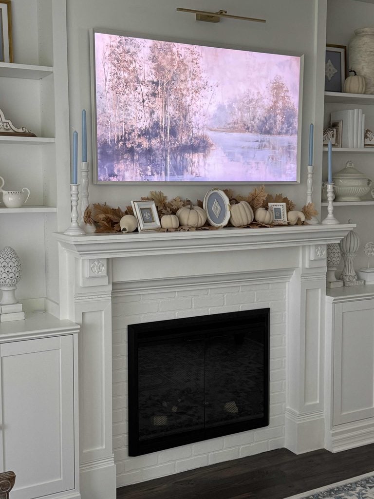 White fireplace with a mantel decorated with white pumpkins, dried leaves, framed art, and two blue candles. Above is a large framed landscape painting of trees by a lake. Shelves with white decor flank the fireplace.