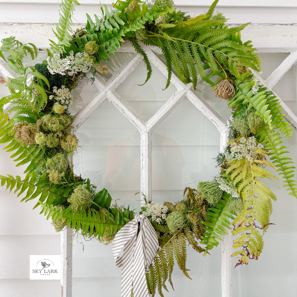 A decorative wreath made of green ferns, moss, and small white flowers hangs on a window. A striped fabric ribbon is tied at the bottom. The background shows white-painted window panes.