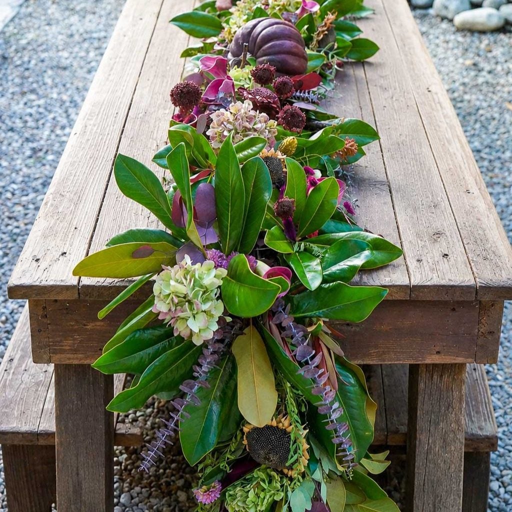A rustic wooden table is decorated with a lush garland of green leaves, hydrangeas, purple flowers, and a striking pumpkin as the centerpiece. The garland cascades elegantly over the table edge.