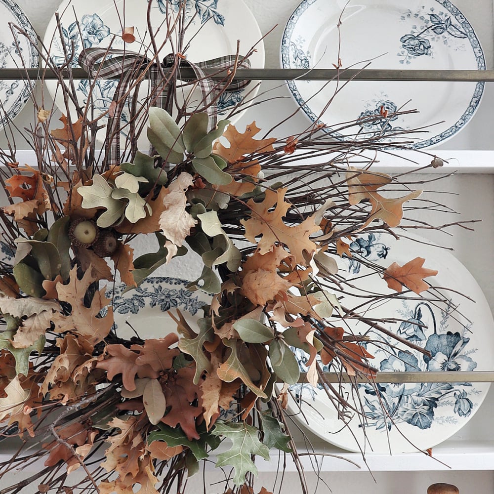 A wreath made of dried leaves, acorns, and twigs hangs in front of white plates with blue floral designs on a shelf, creating a cozy autumn display.