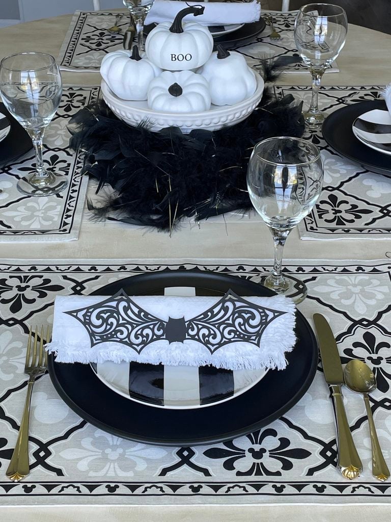 Elegant Halloween table setting with black and white pumpkins, BOO sign, gold cutlery, and a bat napkin holder—perfectly complementing your Halloween mantel decor with chic feather accents and patterned placemats.