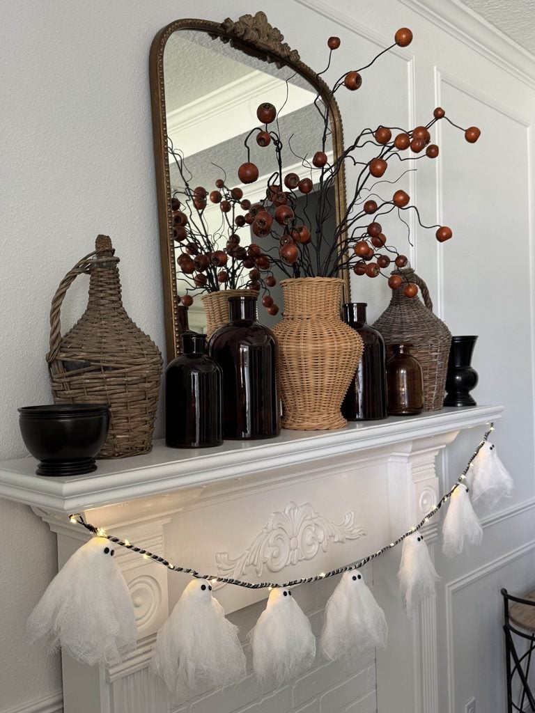Decorative mantel with brown glass and wicker vases, a black bowl, autumn branches, and a large mirror. A DIY ghost garland with small lights hangs across the white ornate fireplace for a festive touch.