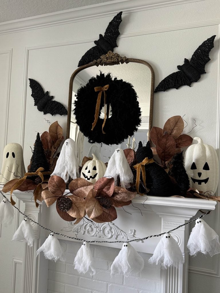 A Halloween mantel decor scene with white ghost and pumpkin figures, black bats on the wall, a wreath with a bow on the mirror, brown leaves, and a garland of white ghosts draped across the front.