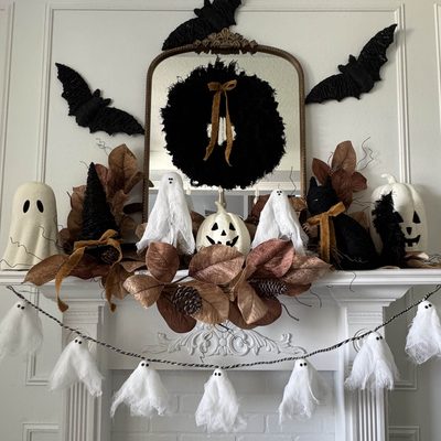 White fireplace mantel featuring Halloween mantel decor with faux leaves, ghost and pumpkin figures, black bats on the wall, a round wreath with a bow, and a hanging ghost garland for a festive display.
