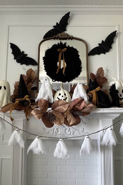 White fireplace mantel featuring Halloween mantel decor with faux leaves, ghost and pumpkin figures, black bats on the wall, a round wreath with a bow, and a hanging ghost garland for a festive display.