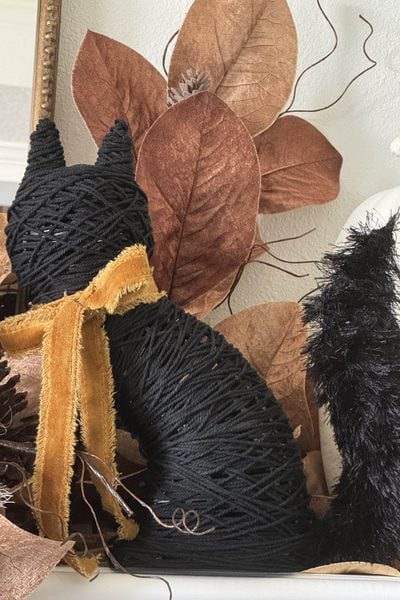 A DIY black cat figure made from wrapped twine with a brown ribbon sits among brown leaves.