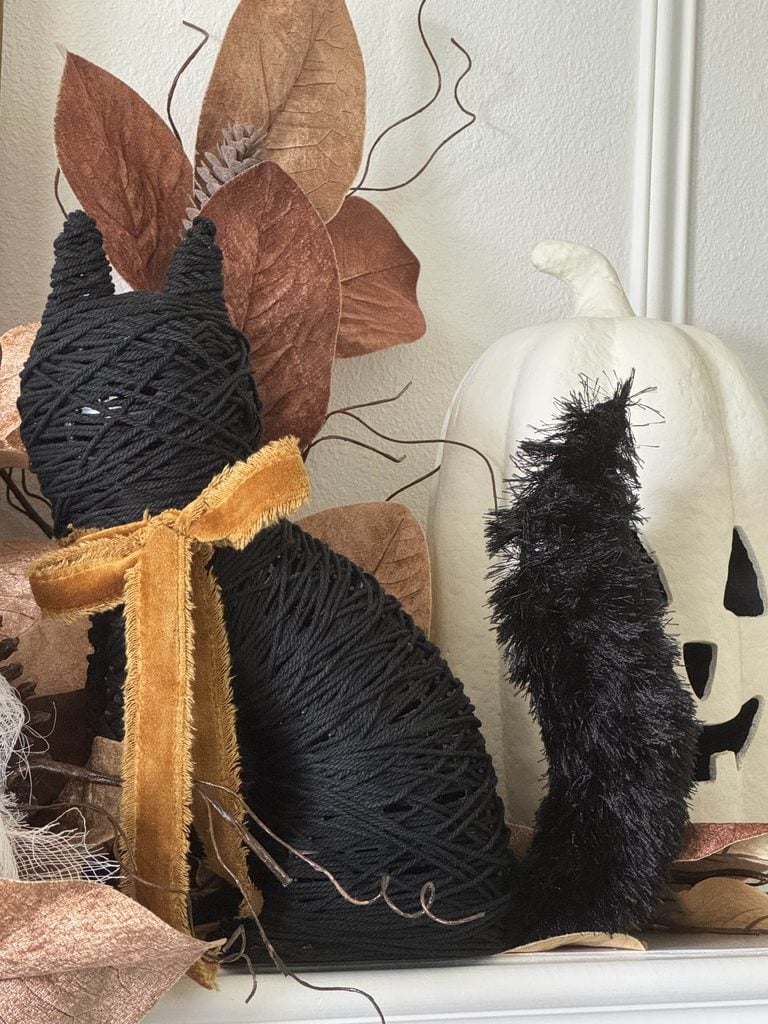 A black yarn-wrapped cat figurine with a gold ribbon sitting next to a white jack-o-lantern, displayed near a DIY ghost garland.